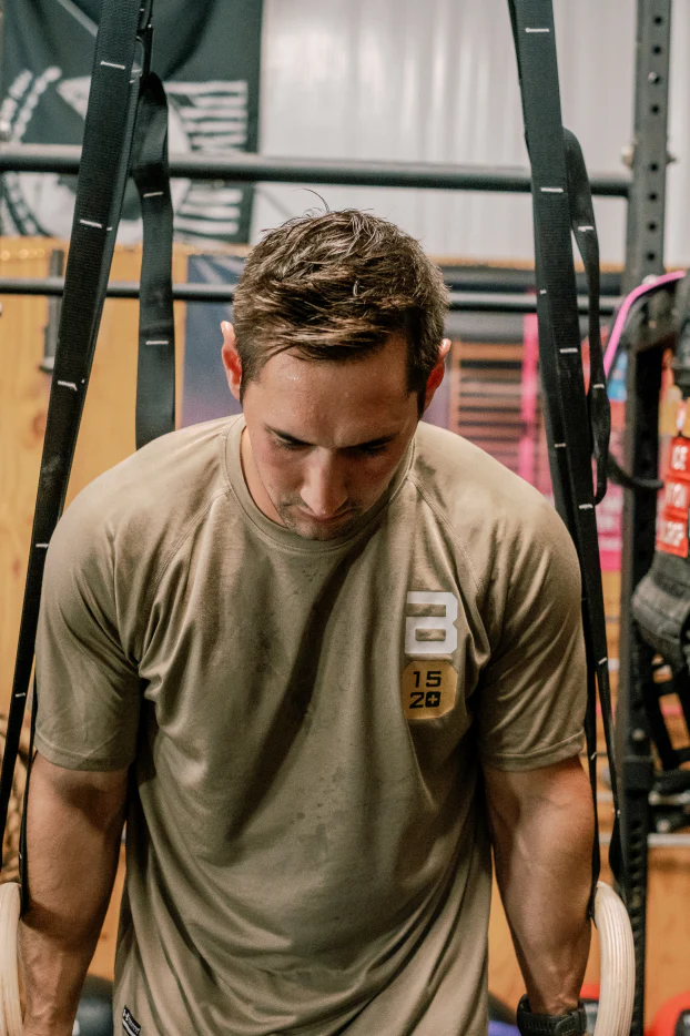 A man in a beige workout shirt using gymnastic rings for strength training in a gym.